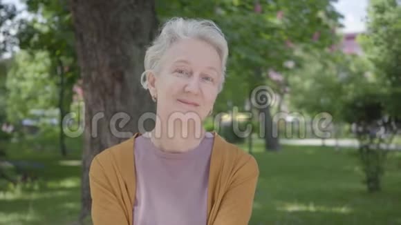美丽的老妇人的肖像灰色的头发和蓝色的眼睛在绿色惊人的公园里微笑成熟的视频的预览图