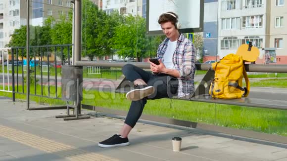 快乐的年轻人正在公共汽车站等车坐在长凳上的男性他用智能手机听音乐视频的预览图