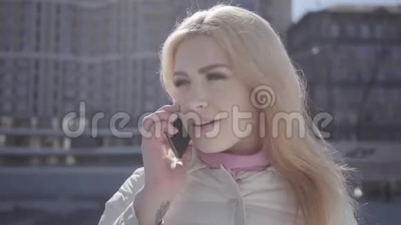 一幅美丽的金发女人的肖像戴着温暖外套的牙套站在城市的街道上用手机交谈城市视频的预览图