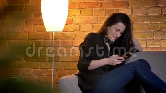 年轻的黑发白种人女孩坐在沙发上在家庭氛围中严肃地制作自拍照片的肖像视频的预览图