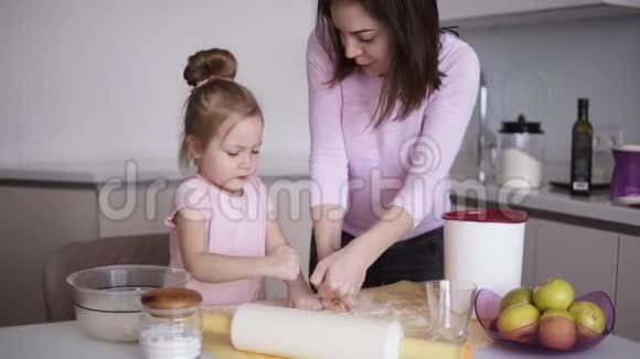 漂亮的年轻妈妈和她可爱的小女儿正在家里用手在厨房里制作桌子上的面团慢慢视频的预览图