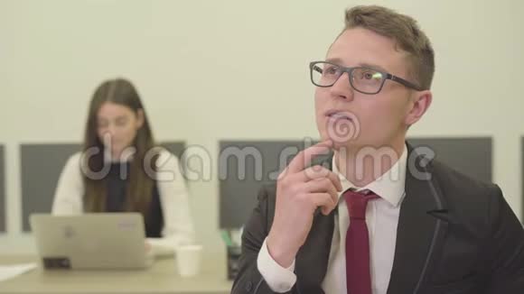 肖像英俊体贴的年轻人戴着眼镜坐在办公室的前台而他的女同事则在视频的预览图