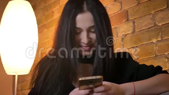 年轻的黑发白种人女孩在智能手机上打字的肖像在家庭氛围中欣然地微笑着进入相机视频的预览图