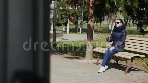年轻女子在阳光明媚的公园里的长椅上喝咖啡视频的预览图