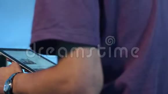 穿紫色t恤的苗条男性坐在黑暗的房间里积极使用他的平板电脑视频的预览图