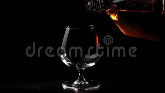 豪华白兰地在黑色背景下旋转的黑色桌子上用手将圆形瓶子中的金色干邑倒入玻璃杯中布兰迪视频的预览图