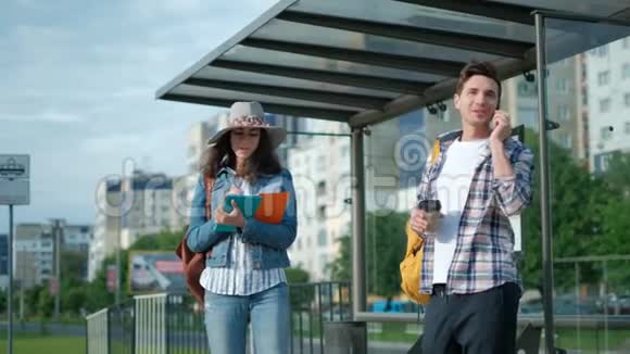 年轻人和女孩站在公共汽车站太阳照耀着他在用手机在等他的电车城市视频的预览图