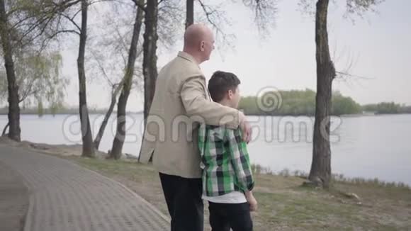爷爷和孙子站在河岸上望着水面那个男人用肩膀拥抱孩子视频的预览图