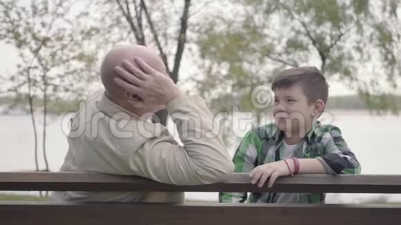 爷爷和孙子坐在河边的公园里的长凳上老人给男孩讲了一个有趣的故事视频的预览图
