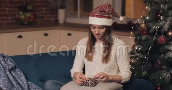 年轻漂亮的女人戴着桑塔帽坐在沙发上在舒适的圣诞节背景下拿着小礼物盒视频的预览图