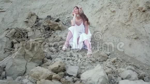 美丽的年轻女孩穿着白色的长裙头上戴着围巾拖鞋坐着想念悬崖附近的石头他打招呼视频的预览图
