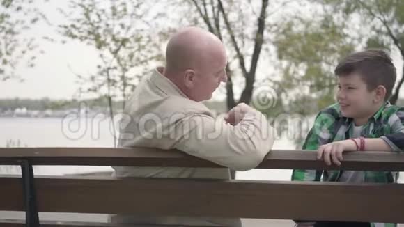 爷爷和孙子坐在河边的公园里的长凳上老人给男孩讲了一个有趣的故事视频的预览图