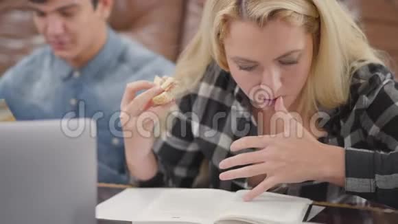 肖像可爱的金发女孩一边看笔记本一边吃三明治而她的男朋友在网上搜索信息视频的预览图