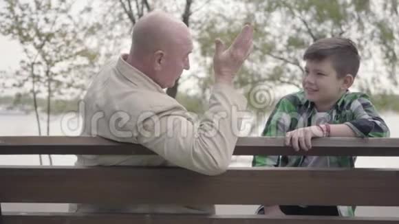 肖像祖父和孙子坐在河边的公园里的长凳上老人讲着有趣的故事给人听视频的预览图