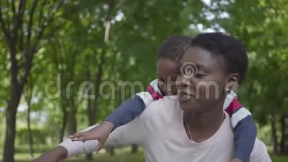 非洲裔美国妇女抱着儿子背在绿色公园的肖像手臂张开紧贴着侧面可爱视频的预览图