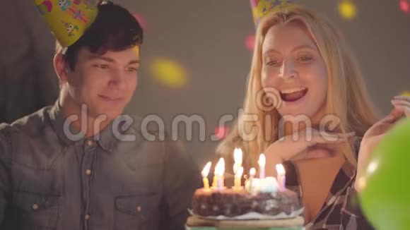 可爱的微笑金发女孩戴着生日帽吹灭蛋糕上的蜡烛年轻人坐在附近女人有视频的预览图
