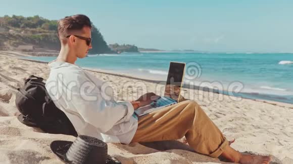 千禧一代年轻的城市数字游牧者坐在海滩上用笔记本电脑工作视频的预览图
