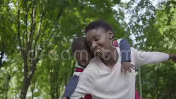 可爱的非洲裔美国妇女抱着儿子背在绿色公园的肖像手臂张开紧贴着侧面视频的预览图