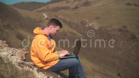 一个穿着黄色夹克蓝色牛仔裤和眼镜的人坐在山上为笔记本电脑工作紧张发誓喊叫视频的预览图