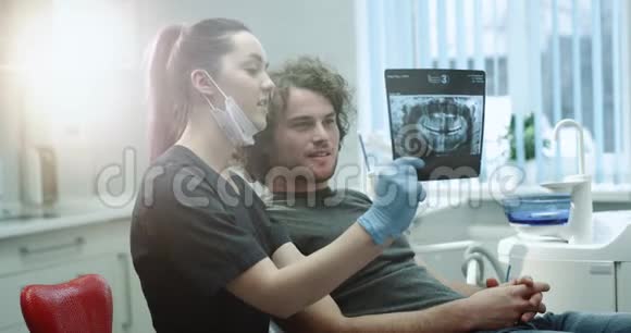 带着蓝色手套和口罩的年轻牙医向病人解释他的牙齿X光检查结果并向男子展示他的牙齿X光检查结果视频的预览图