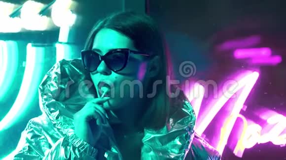 年轻的时尚少女戴着毛皮眼镜舔着一颗糖果上面有霓虹灯蓝色粉红色的标志美丽的千禧年视频的预览图