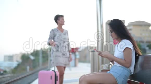 年轻的亚洲妇女在火车站用电话与成熟的旅游妇女问路视频的预览图