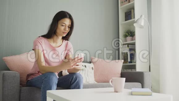漂亮的年轻女人在家里坐在沙发上用智能手机打字视频的预览图