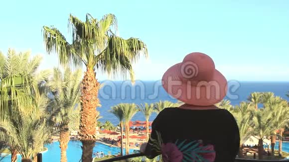 一个阳光明媚的夏日戴着帽子的年轻美女从阳台望向蔚蓝的大海和棕榈树视频的预览图