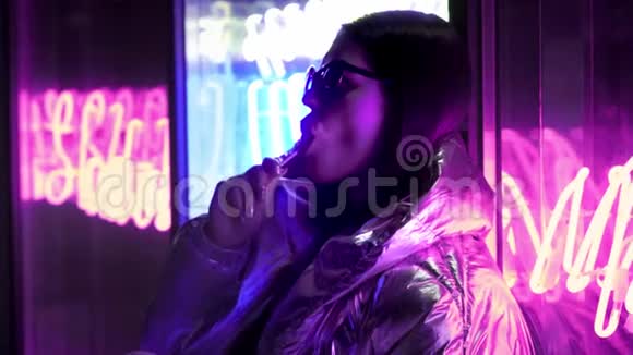 年轻的时尚少女戴着毛皮眼镜舔着一颗糖果上面有霓虹灯蓝色粉红色的标志美丽的千禧年视频的预览图