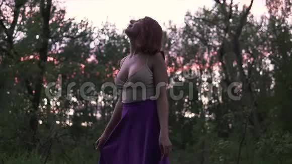 黄昏时分年轻漂亮的幸福女人胸脯大光着脚在树林里转穿着紫色的裙子视频的预览图