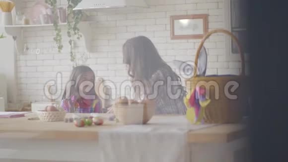 漂亮的妈妈和可爱的女儿在厨房里拿着篮子和复活节彩蛋坐在复活节餐桌上聊天视频的预览图