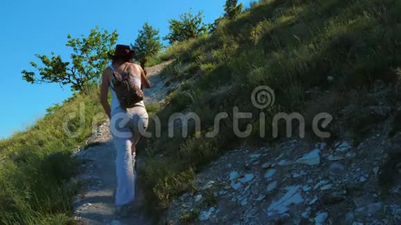 一个带着背包和木棍的女孩旅行者正沿着一条位于山上陡峭斜坡上的小路走着视频的预览图