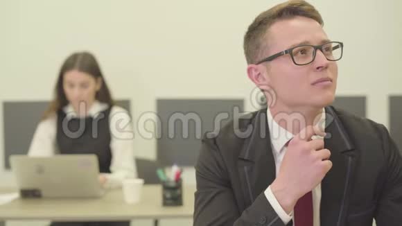 肖像英俊体贴的年轻人戴着眼镜坐在办公室的前台而他的女同事则在视频的预览图