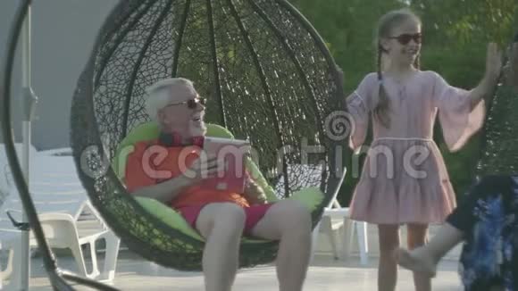一对老夫妇坐在一张挂椅上一起在酒店综合体放松可爱的小女孩站在附近视频的预览图