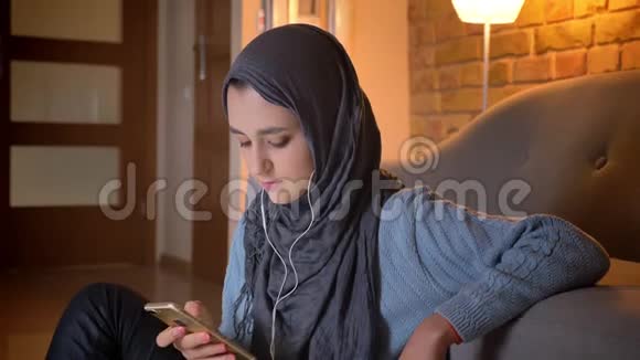 年轻迷人的穆斯林女性在坐在地板上时使用听音乐的手机进行震动特写拍摄视频的预览图