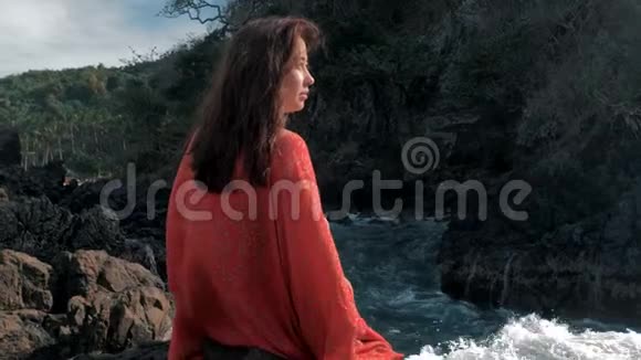 年轻女孩穿着红色衣服坐在暴风雨的附近海浪拍打着岩石年轻女孩望着大海海浪拍打着视频的预览图