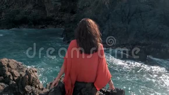 年轻女孩穿着红色衣服坐在暴风雨的附近海浪拍打着岩石年轻女孩望着大海海浪拍打着视频的预览图