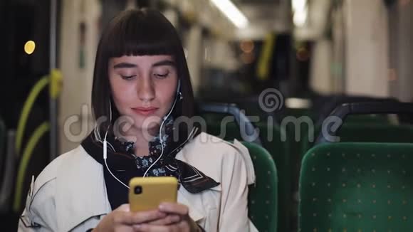 年轻时尚的女士戴着耳机听音乐在公共交通工具上浏览手机城市视频的预览图