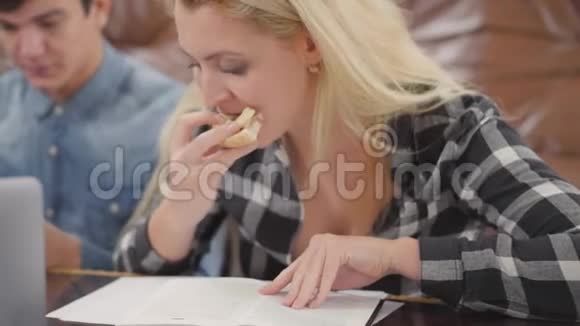 那个金发女孩在看笔记本时吃三明治而她的男朋友则用笔记本电脑在网上搜索信息视频的预览图