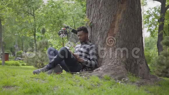 英俊的非裔美国人坐在公园的一棵老树下的自行车旁看书户外休闲视频的预览图