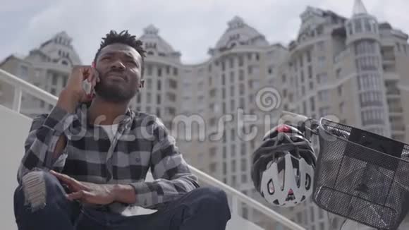 一个英俊的非裔美国人坐在自行车旁以城市建筑为背景说话的人视频的预览图