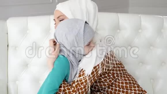 穆斯林妈妈和她戴着头巾的小女儿抱着坐在沙发上视频的预览图