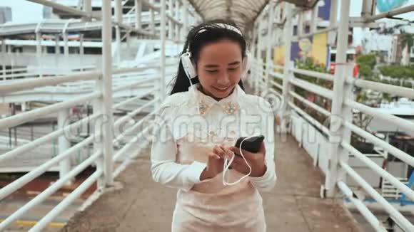 一个开朗的印尼女孩站在城市的一座桥上戴着白色耳机在智能手机上选择音乐视频的预览图