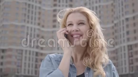 在摩天大楼前用手机说话的可爱微笑自信的金发女人的特写肖像城市生活方式视频的预览图