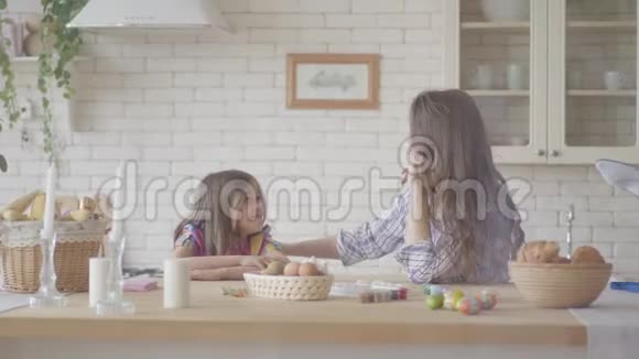 可爱的母亲和漂亮的女儿在厨房里拿着篮子和复活节彩蛋坐在复活节餐桌上聊天视频的预览图