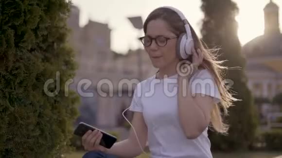 有趣的年轻学生坐在公园的草地上用大耳机听音乐女孩唱歌视频的预览图