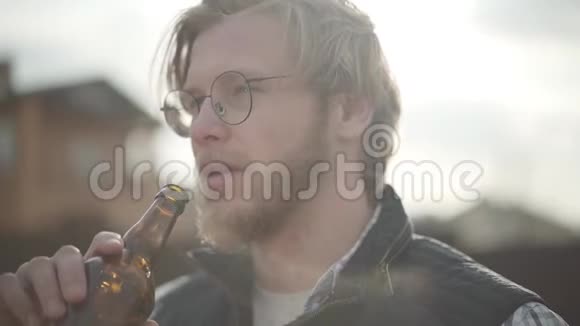 一幅金发男人在户外喝啤酒的肖像戴眼镜的长胡子男人享受他的酒精饮料头发视频的预览图
