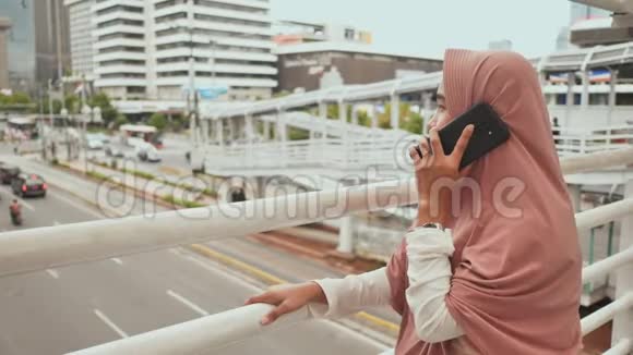 一个穿着粉色头巾的年轻穆斯林女孩在市中心的道路交通上方打电话视频的预览图