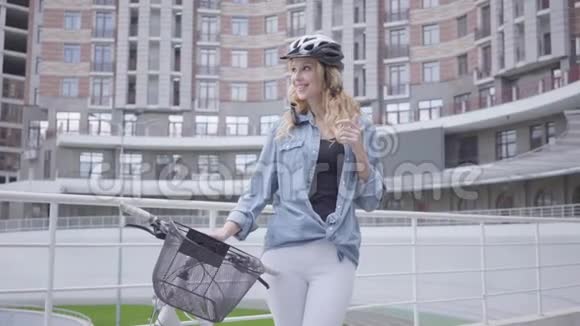 戴着自行车头盔的漂亮金发女人站在城市建筑的背景下骑着自行车女士在忙视频的预览图
