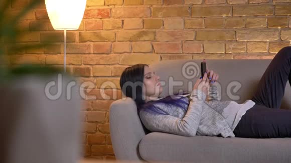 在舒适的家中躺在沙发上用手机浏览年轻漂亮时髦少女的特写镜头视频的预览图